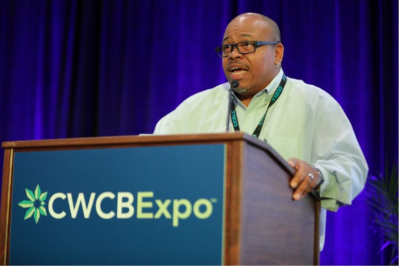 A photograph of Calvin Frye speaking at the podium at CWCBExpo. Calvin is wearing a green shirt and glasses. He is mid-speech.